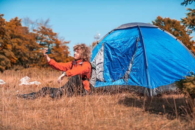 A man with a tent enjoys a sunny day whileusing a smartphone and camping in the mountains