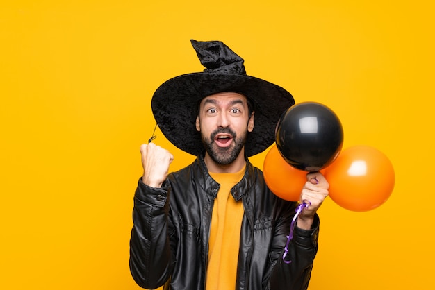 Man with witch hat holding black and orange air balloons for halloween party celebrating a victory in winner position