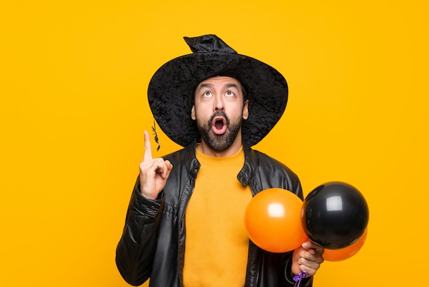 Man with witch hat holding black and orange air balloons for halloween party pointing up and surprised