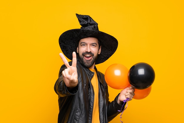 Man with witch hat holding black and orange air balloons for halloween party smiling and showing victory sign