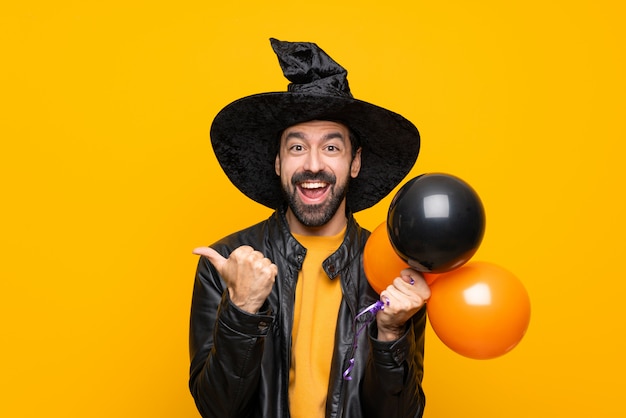 Man with witch hat holding black and orange air balloons for halloween party with thumbs up gesture and smiling