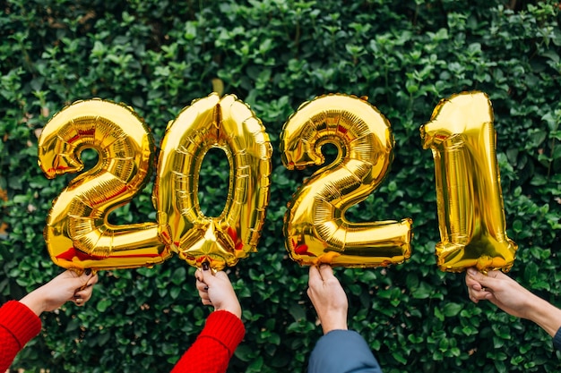 Man and woman couple hands holding gold foil balloons numeral 2021 in front of a wall of plants. New year celebration concept.