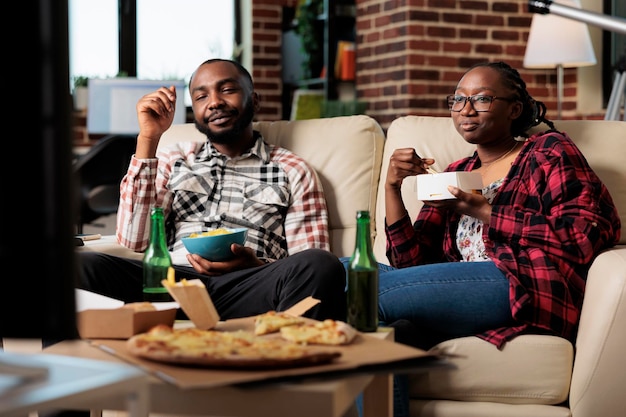 Man and woman enjoying fast food delivery with movie on tv, having fun with leisure activity. Eating takeaway meal with noodles, pizza and chips, drinking beer from bottle. Watching television.