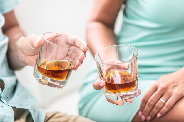 Man and woman hands toasting with glasses of whiskey brandy or rum indoors - closeup.