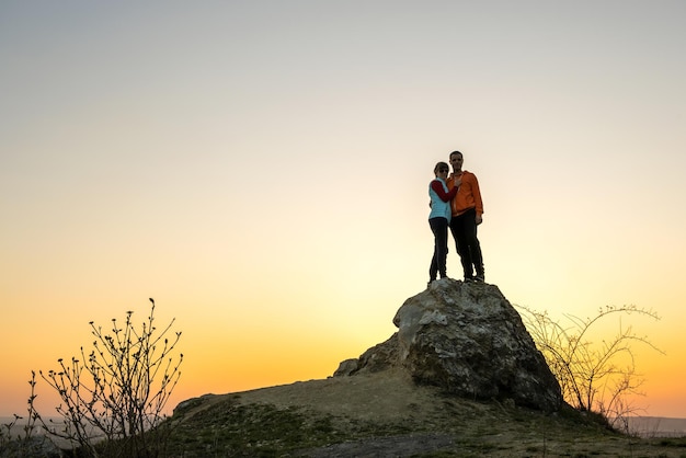 Man and woman hikers standing on a big stone at sunset in mountains Couple together on a high rock in evening nature Tourism traveling and healthy lifestyle concept