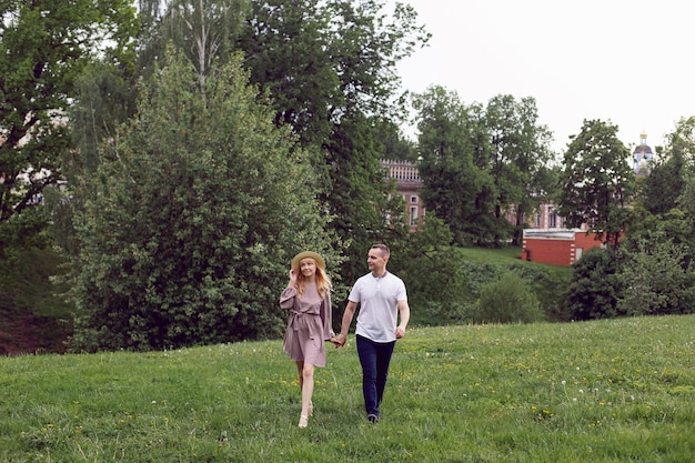 Man and a woman in love in a dress and hat are walk on a green field under a tree in summer