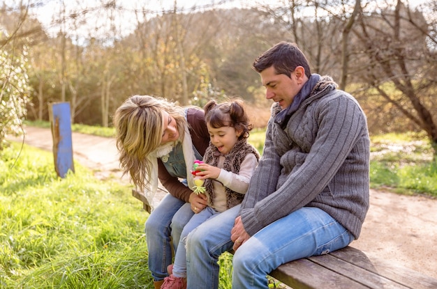 Man and woman playing with a little girl sitting on the center of a wooden bench in the park. Family leisure outdoors concept.