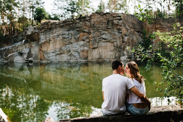 Man and woman sit back, kissing on stone near lake on surface of big rocks