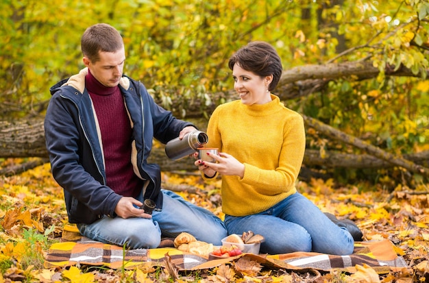 A man and a woman sit on a plaid in an autumn park and drink a hot drink from a thermos