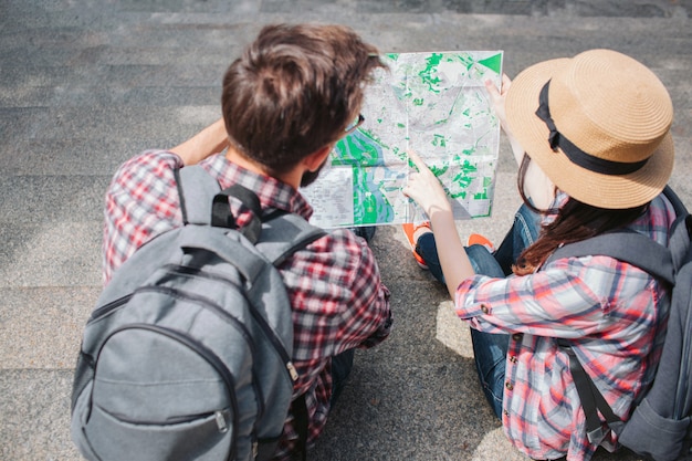 Man and woman sits on steps and holds map together. They look at it. Tourists have rocksacks on back. Woman wears hat.