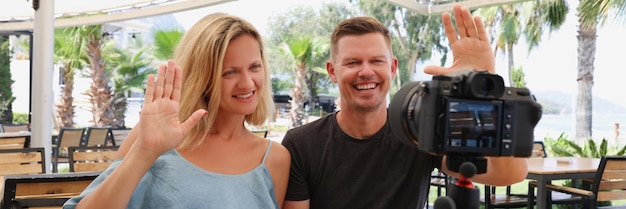 A man and a woman wave to the camera on a tripod in a street cafe travel vlog family vacation