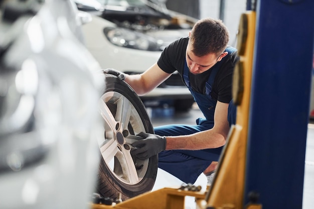 Man in work uniform sitting and changing car wheel indoors Conception of automobile service