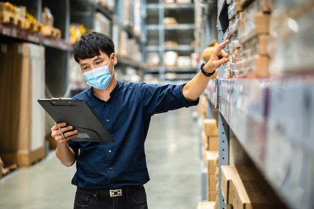 Man worker with medical mask holding clipboard and checking inventory in the warehouse during coronavirus pandemic