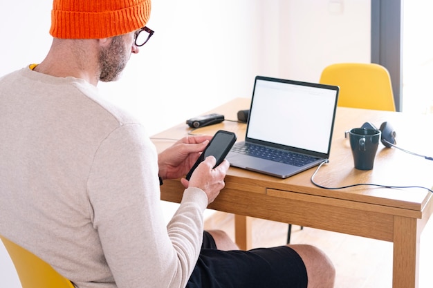 Man working from home on a laptop and smart phone sitting at a desk surfing the internet