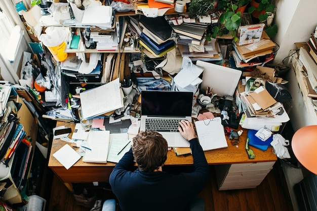 Photo a man works on a laptop at a cluttered desk with a child beside him surrounded