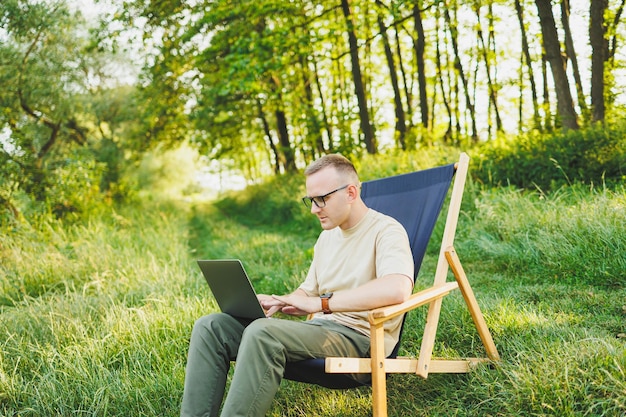 A man works on a laptop while sitting on a bench in a city park A young guy reads the news writes a message on his laptop Recreation in nature remote work on vacation