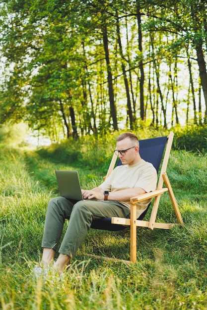 A man works on a laptop while sitting on a bench in a city park A young guy reads the news writes a message on his laptop Recreation in nature remote work on vacation