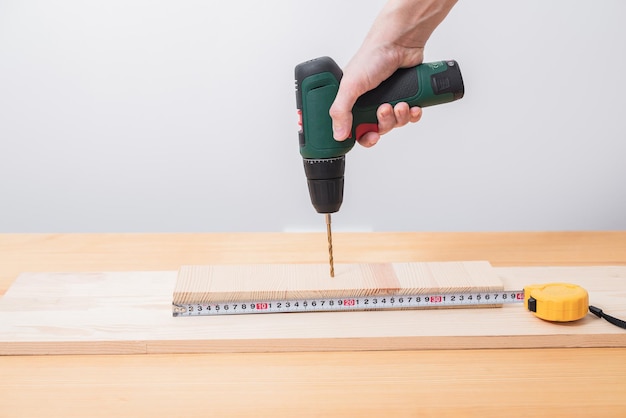 A man works with an electric screwdriver on a wooden table also makes measurements with a tape measure