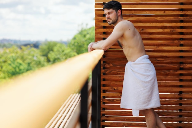 Man wrapped in a towel stands on the balcony of a spa resort leaning against the railing