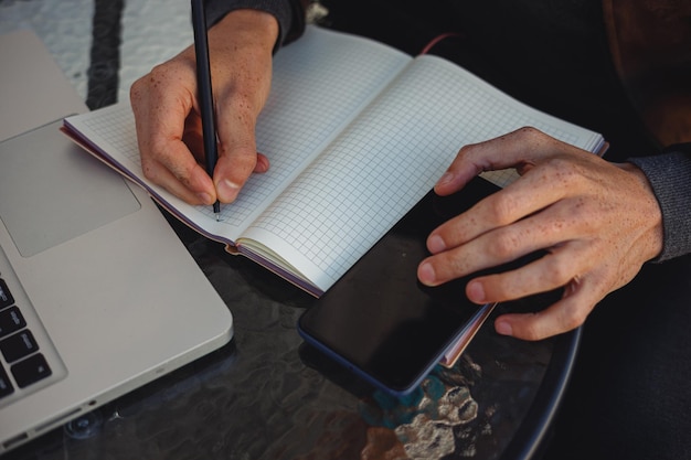 man writing in notepad and making notes while using mobile phone during work in cafe