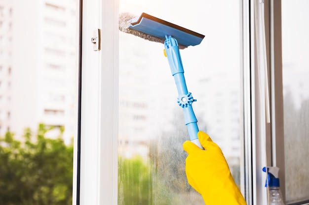 Man in yellow gloves cleaning window with squeegee and spray detergent at home terrace House cleaning and house chores domestic hygiene Window cleaning background with blue sky