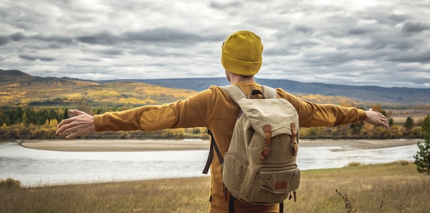 Man in a yellow hat and sweater with a backpack is standing with his arms outstretched to the sides in front of the river, the autumn golden forest and the hills. Concept of freedom, travel, hiking