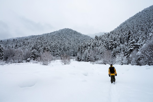 A man in a yellow jacket and backpack walks along a snowcovered path to the mountains Beautiful landscape in Arkhyz with mountains snow and forest on a cloudy winter day Caucasus Mountains Russia