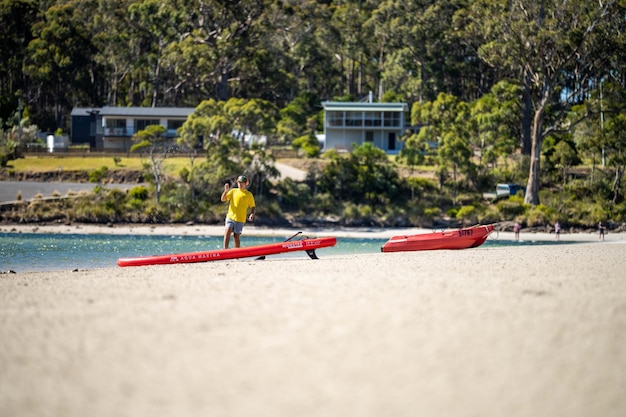 A man in a yellow shirt stands on a beach next to a red kayak.