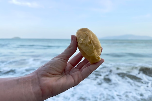 A man39s hand holds a piece of badsmelling ripe durian against the background of the sea ocean water in a tropical country