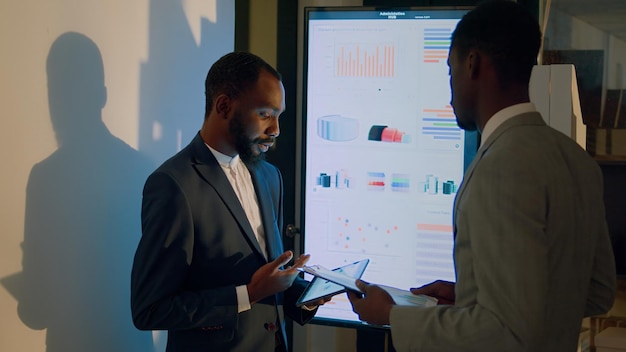 Management executive debriefing worker in front of digital screen, showing him business data on tablet. Company personnel in office during nightshift crosschecking analytical datasets