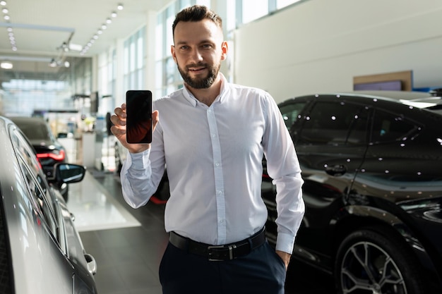 Manager of a car dealership among new cars demonstrates the screen of a smartphone