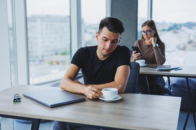 Manager employee sits at a wooden table and works on a laptop There is coffee and a laptop on the table Office with large windows