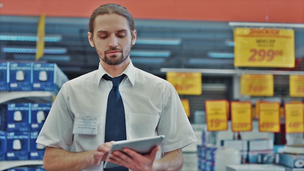 Photo manager man doing stocktaking of products in cardboard box on shelves in warehouse using digital tablet and pen male professional assistant checking stock in factory physical inventory count