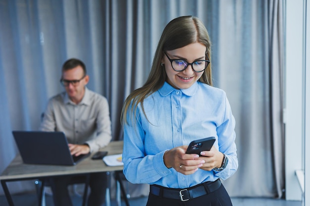 Manager woman in classic glasses smiling during working hours in the office talking on the phone table with laptop colleagues in the background A colleague is in the background selective focus