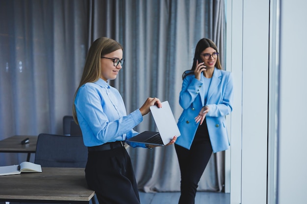 Manager woman in classic glasses smiling during working time in office positive european woman in blue shirt desk with laptop remote work A colleague is in the background selective focus