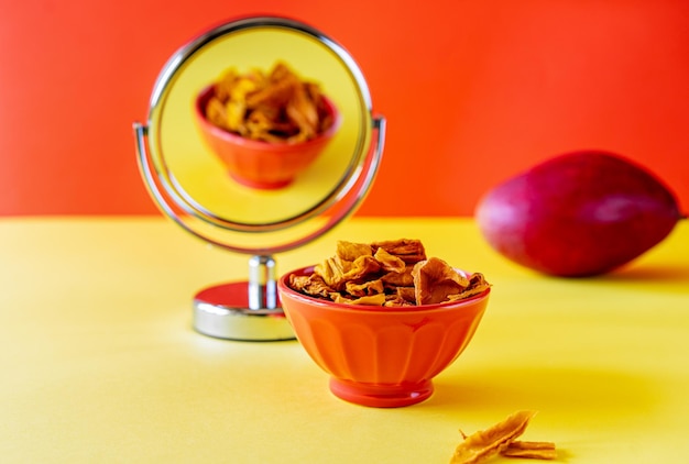 Mango chips in bowl with mirror and raw mango over yellow and orange background