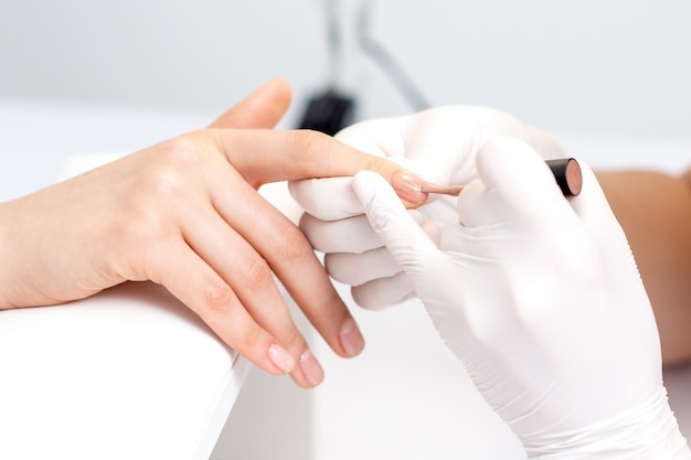 Photo manicure master in protective gloves applying beige nail polish on female nails in beauty salon