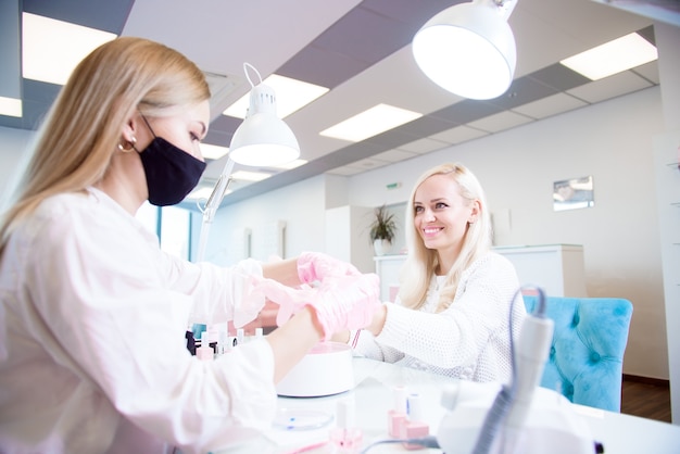 Manicure and pedicure salon, covid-19 and social distance. Master in rubber gloves and young woman client in protective mask in beauty studio interior.