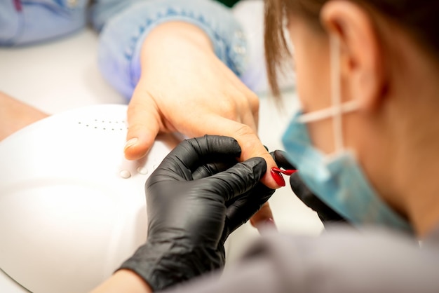 Manicure varnish painting Closeup of a manicure master wearing rubber black gloves applying red varnish on a female fingernail in the beauty salon