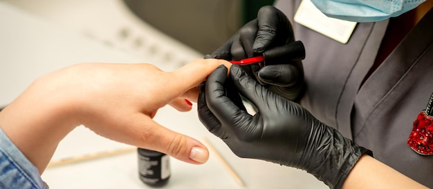 Manicure varnish painting Closeup of a manicure master wearing rubber black gloves applying red varnish on a female fingernail in the beauty salon