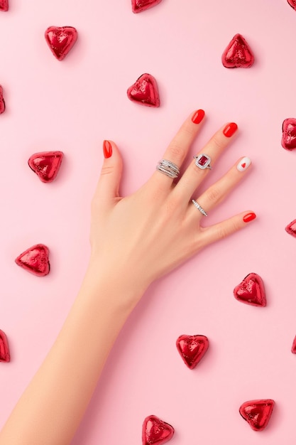 Manicured womans hand with rings on pink background fashionable red nail design with heart