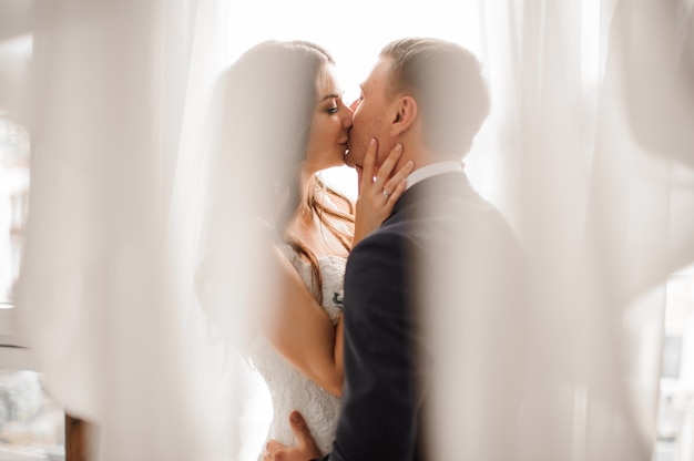 Manly groom and beautiful bride kissing against white scene