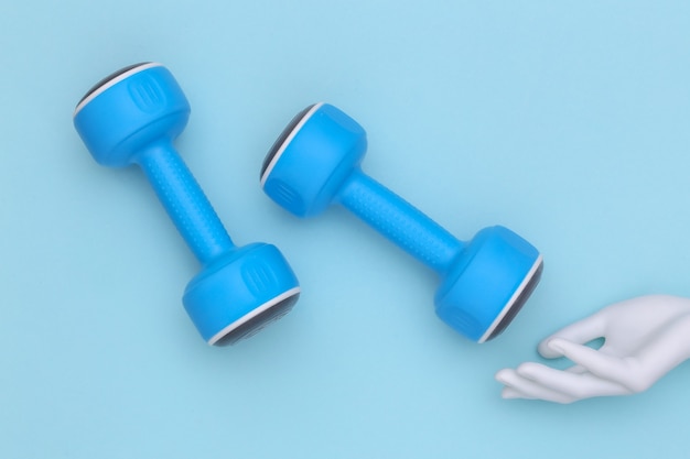 The mannequin's white hand touches plastic dumbbells on blue background. Sport and fitness concept