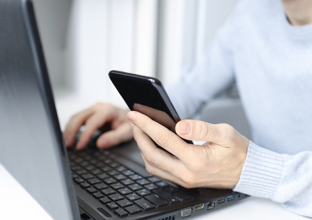 A mans hand holding a smartphone working on a laptop