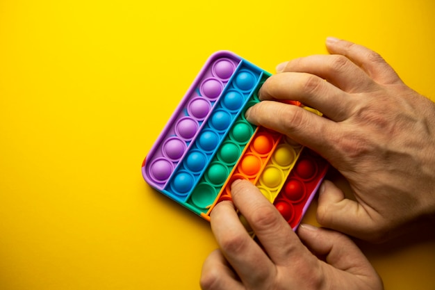 Photo a mans hand presses on a rainbow silicone antistress toy pop it or a simple dimple on a yellow background copy space