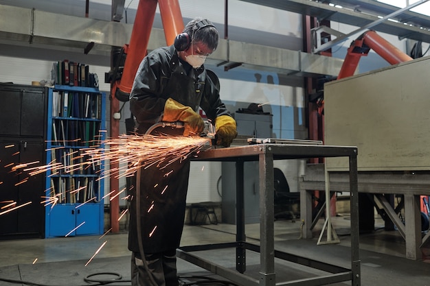 Manual worker in mask and ear protectors standing at high metal table and cutting metal with rotary tool in industrial shop