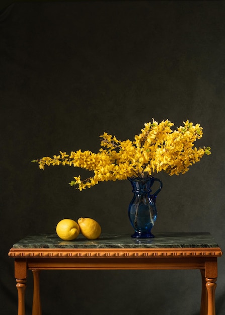 Many branches with yellow flowers in a blue glass vase jug on a gray background A bouquet of yellow flowers on a marble table is lined with two lemons Vitamin C Good health