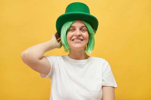 March festivities in Ireland Celebrate Irish traditions Beautiful woman wearing festive green hat standing isolated over yellow background looking at camera with toothy smile