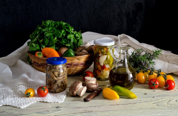 Marinated vegetables and mushrooms in jars and raw cherry tomatoes peppers parsley and mushrooms in a basket on a wooden table closeup