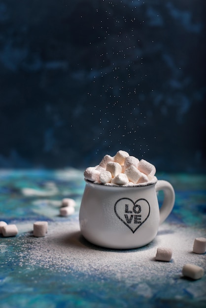 Marshmallows in cocoa in white mug with scattered mini marshmallows on blue